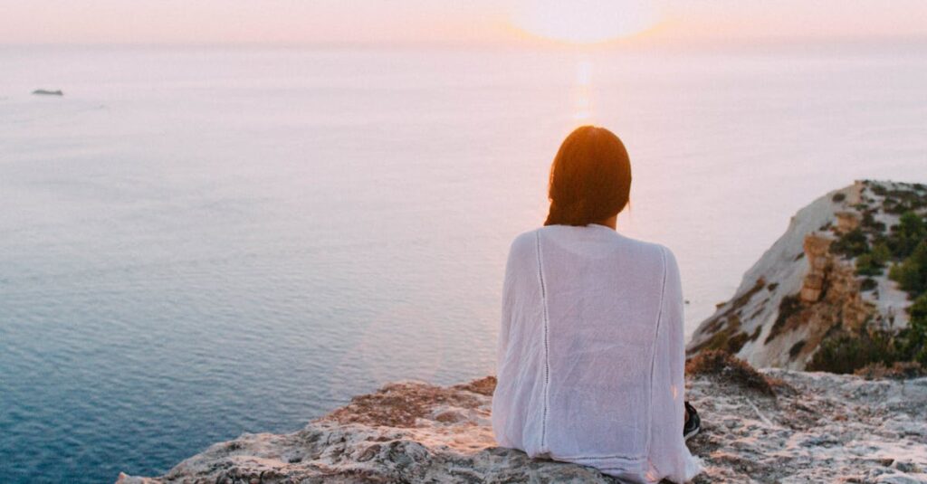 A serene view of a woman sitting on a rocky cliff, gazing at the ocean during sunset. Perfect for relaxation and travel themes.