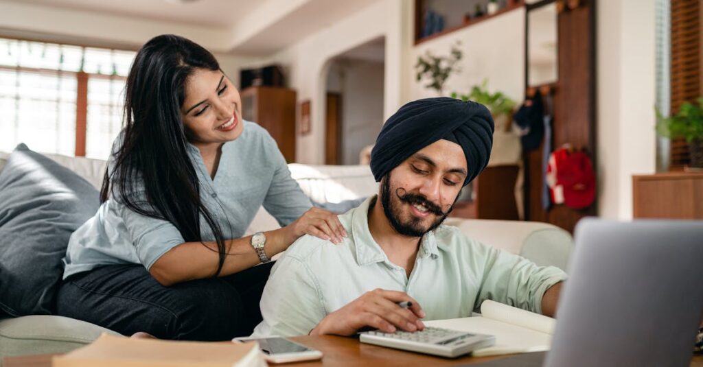 Positive ethnic man sitting at coffee table with laptop and smartphone and books and notebook and calculating while smiling ethnic wife sitting on sofa and supporting and watching behind