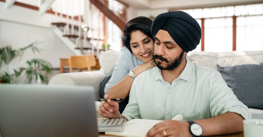 Cheerful young Indian woman cuddling and supporting serious husband working at home with laptop and counting on calculator